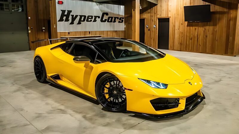 A yellow Lamborghini sports car with black accents is parked indoors near a wooden wall with a "HyperCars" banner—one of the best supercars under $200k and currently for sale.