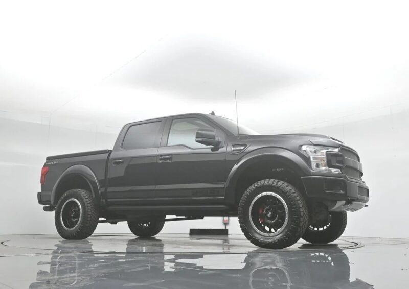 A black Shelby Trucks four-door pickup with large off-road tires is parked in a brightly lit, white indoor studio.