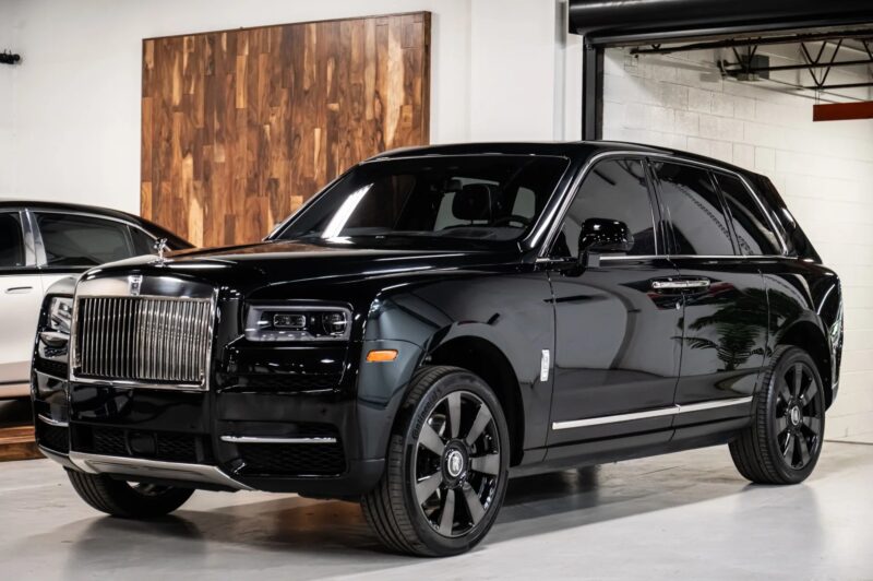A black Rolls-Royce Cullinan luxury SUV is parked indoors on a concrete floor near a white wall and a wooden panel, showcasing elegance and style.