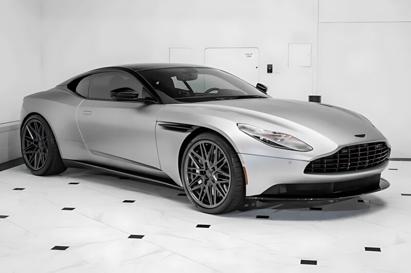 A silver Aston Martin coupe with a black roof and large alloy wheels is parked indoors on a white floor with black accents—for sale: one of the best supercars under $200k.