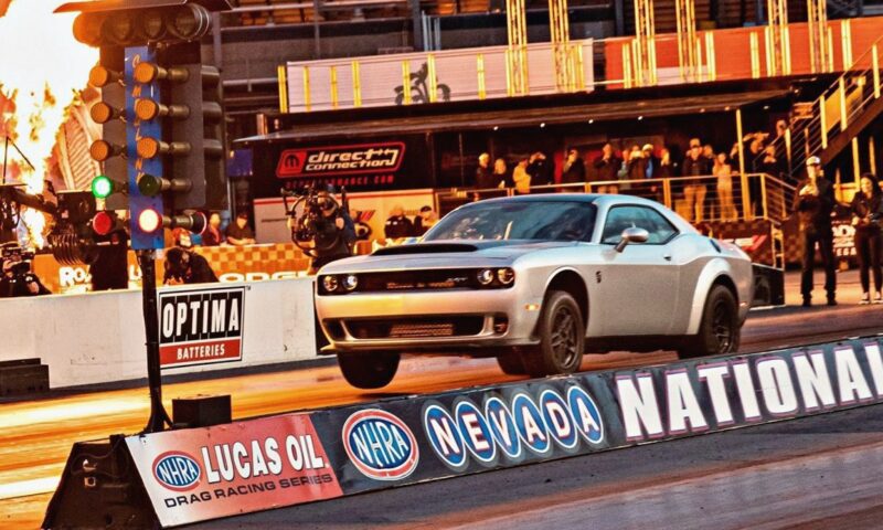 A silver 2023 Dodge Challenger SRT Demon 170 drag races on the track, its front wheels lifted as fire blazes in the background. The crowd watches and NHRA signage is visible—a crown jewel no MOPAR collector can ignore.