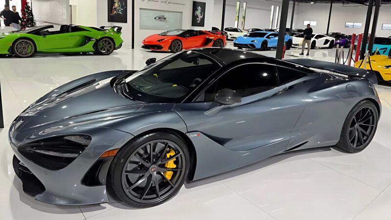 A metallic gray sports car with yellow brake calipers is displayed in a showroom, surrounded by other brightly colored sports cars and marked as one of the best supercars under $200k for sale.