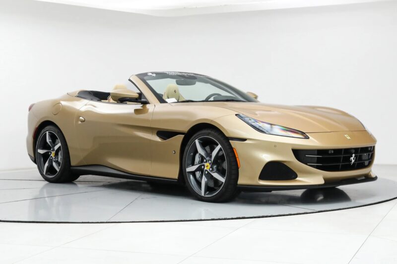 A luxury car, this gold Ferrari Portofino convertible is parked in a white indoor studio with its top down, shown from a front-side angle.