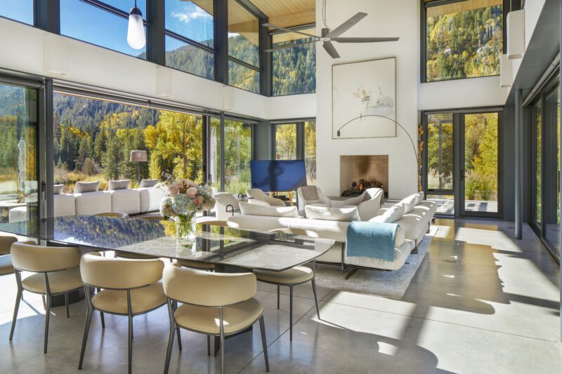 Modern living and dining area with large windows, neutral furniture, a glass dining table, fireplace, and scenic views that capture the essence of Outdoor Mountain Living in Aspen's forested landscape.