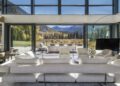 Modern living room with white furniture and large windows, embracing Outdoor Mountain Living and overlooking a scenic Aspen landscape with trees and a patio.