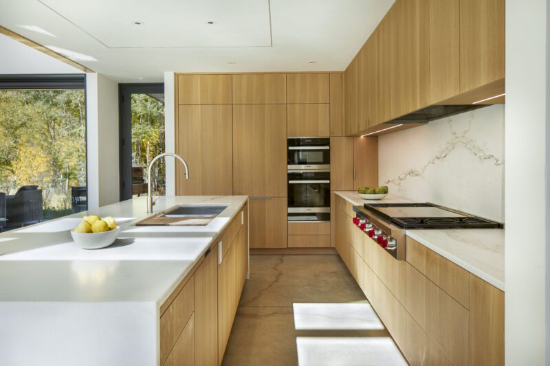 Modern kitchen with light wood cabinets, white countertops, built-in ovens, gas stove, bowl of lemons, and a large window showcasing outdoor greenery—perfect for embracing the Outdoor Mountain Living lifestyle in Aspen.