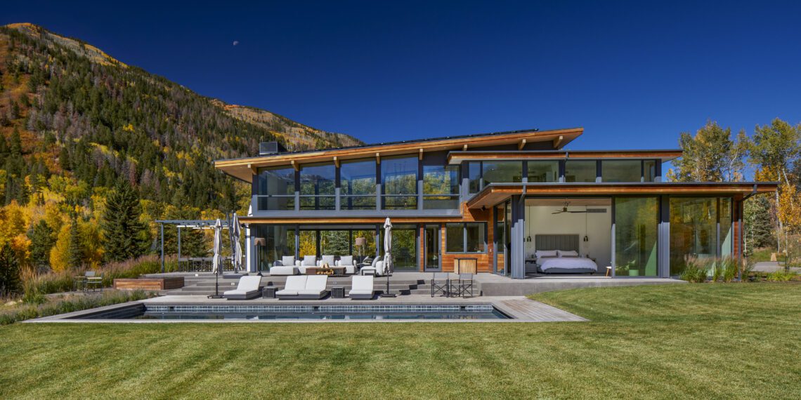 Modern glass-walled house with outdoor seating and a pool, set against a backdrop of green lawn, trees, and a mountain under a clear blue sky.