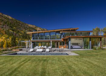 Modern glass-walled house with outdoor seating and a pool, set against a backdrop of green lawn, trees, and a mountain under a clear blue sky.