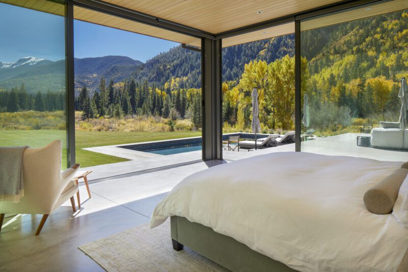Bedroom with large floor-to-ceiling windows overlooking a patio, pool, and scenic Aspen mountain landscape—perfect for embracing outdoor mountain living amid trees in autumn colors.