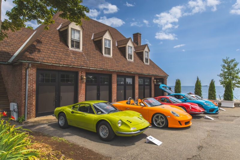 A row of colorful sports cars, including yellow, orange, red, and blue models, parked in front of a brown brick building with four garage doors near a body of water at the Misselwood Concours d’Elegance 15th Anniversary this July.