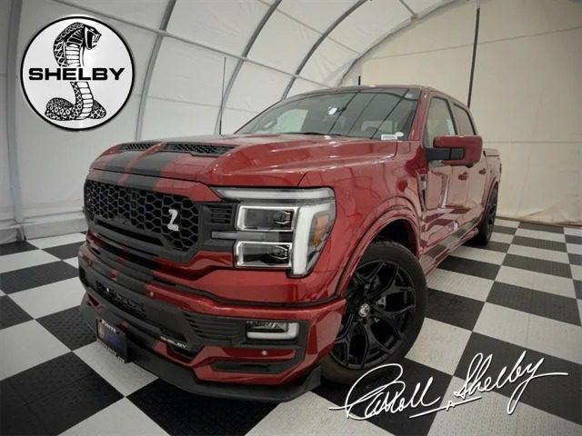 Red Shelby pickup truck with black racing stripes displayed in a white tented showroom, featuring Shelby Trucks logos and Carroll Shelby’s signature on the floor—a must-see for Ford F-150 enthusiasts searching for trucks for sale.