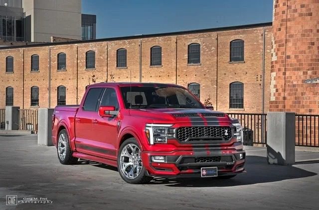 A red Ford F-150 pickup truck with black racing stripes, inspired by Shelby trucks, is parked in an urban setting in front of a brick building—perfect for anyone searching for standout trucks for sale.