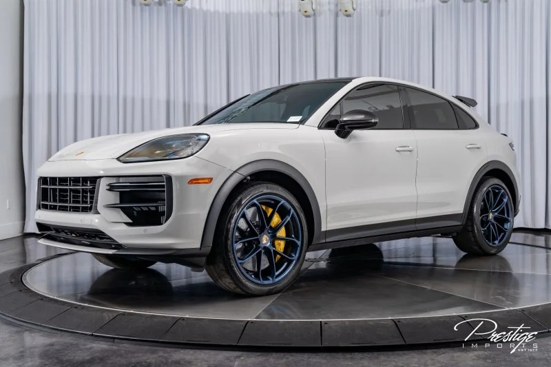 A white Porsche Cayenne luxury car is displayed in a showroom with white curtains and dark flooring, featuring black wheels and yellow brake calipers.