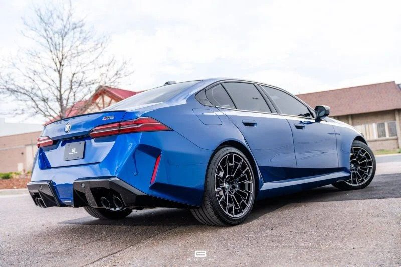 A blue BMW luxury car sedan is parked outdoors on a paved surface, viewed from the rear passenger side with buildings in the background.