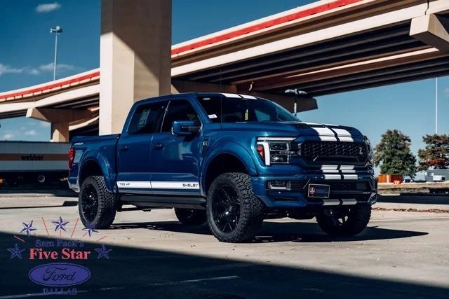 A blue Ford F-150 pickup truck with white racing stripes is parked under an overpass near a sign for Sam Pack’s Five Star Ford Dallas, showcasing one of the standout trucks for sale in the area.