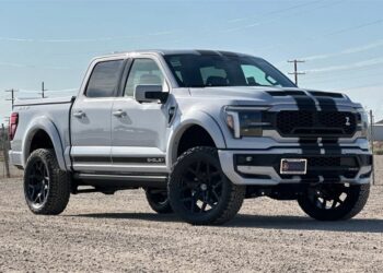 A white Ford F-150 pickup truck with black racing stripes, inspired by Shelby Trucks, is parked on a gravel lot under a clear sky. For sale now—don’t miss your chance to own this striking vehicle!.