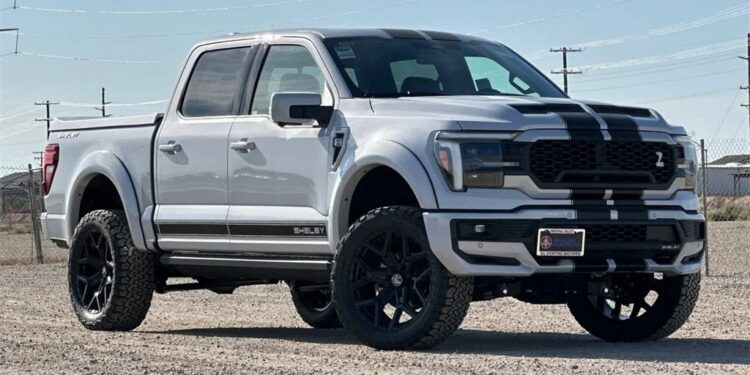 A white Ford F-150 pickup truck with black racing stripes, inspired by Shelby Trucks, is parked on a gravel lot under a clear sky. For sale now—don’t miss your chance to own this striking vehicle!.