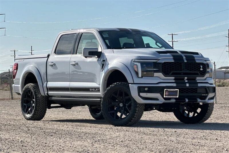 A white Ford F-150 pickup truck with black racing stripes, inspired by Shelby Trucks, is parked on a gravel lot under a clear sky. For sale now—don’t miss your chance to own this striking vehicle!.