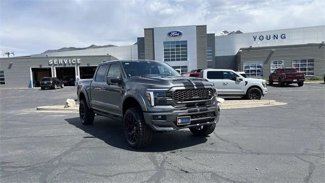 A gray Ford F-150 pickup truck is parked outside a Ford dealership, with other trucks and the dealership building visible in the background and several vehicles for sale nearby.