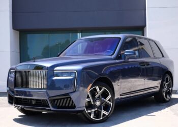 A blue Rolls-Royce Cullinan luxury SUV with a prominent chrome grille, angular headlights, and large alloy wheels is parked in front of a modern building.