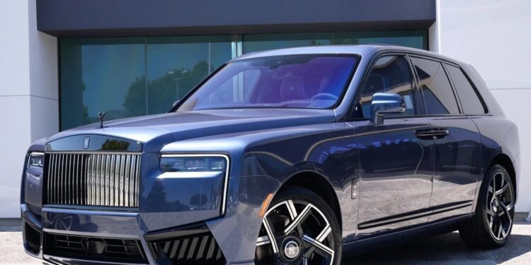 A blue Rolls-Royce Cullinan luxury SUV with a prominent chrome grille, angular headlights, and large alloy wheels is parked in front of a modern building.