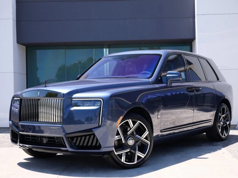 A blue Rolls-Royce Cullinan luxury SUV with a prominent chrome grille, angular headlights, and large alloy wheels is parked in front of a modern building.