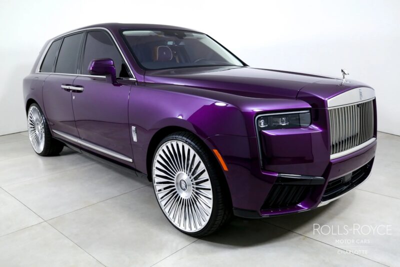 A purple Rolls-Royce Cullinan luxury SUV with large chrome wheels is parked indoors on a light-colored floor, viewed from the front left angle.