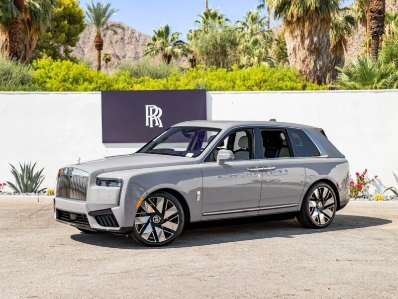A silver Rolls-Royce Cullinan luxury SUV is parked on a street in front of a sign with the Rolls-Royce logo, surrounded by palm trees and other greenery, showcasing the pinnacle of automotive elegance.