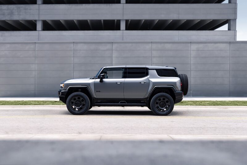 A silver GMC HUMMER EV SUV is parked on the street in front of a multi-level parking garage and a gray concrete wall, showcasing the bold presence of this 2026 electric vehicle.