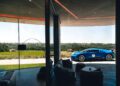 A blue sports car is parked outside a modern home with large glass windows, overlooking a green landscape and distant hills.