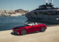 A red 2026 Mercedes-Maybach SL 680 convertible is parked on a marina dock next to a large luxury yacht, with boats and buildings visible in the background. This stunning model will soon be available at US dealers.