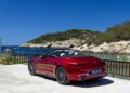 A 2026 Mercedes-Maybach SL 680 convertible car parked by a seaside railing, with clear blue water and rocky hills in the background under a sunny sky. Arriving soon at US dealers.