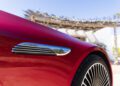 Close-up of a red 2026 Mercedes-Maybach, highlighting the chrome Maybach emblem above the front wheel, with a blurred background of a pergola and building.