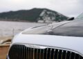 Close-up of the front grille and hood ornament of a 2026 Mercedes-Maybach SL 680, with a body of water and a mountain in the background, soon to be available at select US dealers.