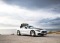 A white 2026 Mercedes-Maybach SL 680 convertible with its black roof partially open is parked on a coastal road next to the sea under a cloudy sky, highlighting luxury soon available at US dealers.