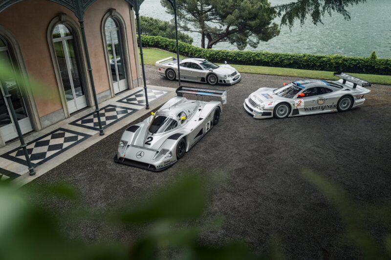 Three silver Mercedes-Benz race cars, including a rare CLK GTR, are parked outside a building with a lake and trees in the background at FuoriConcorso 2025.
