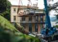 A Mercedes-Benz CLK GTR race car is being lifted by a crane in front of a large house near a lake, with greenery and trees surrounding the FuoriConcorso 2025 scene.
