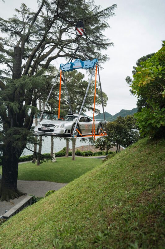 A silver Mercedes-Benz is being lifted by a crane above a grassy hill and trees near a lake, possibly as part of the FuoriConcorso 2025 event.
