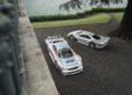 Two silver Mercedes-Benz CLK GTR sports cars are parked on a gravel driveway near a tree and a lake, one featuring racing decals and a large rear wing—an iconic scene from FuoriConcorso 2025.