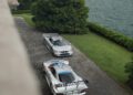 Two silver Mercedes-Benz sports cars, including a rare CLK GTR, are parked on a lakeside driveway surrounded by green trees and mountains under a cloudy sky—an unforgettable scene at FuoriConcorso 2025.