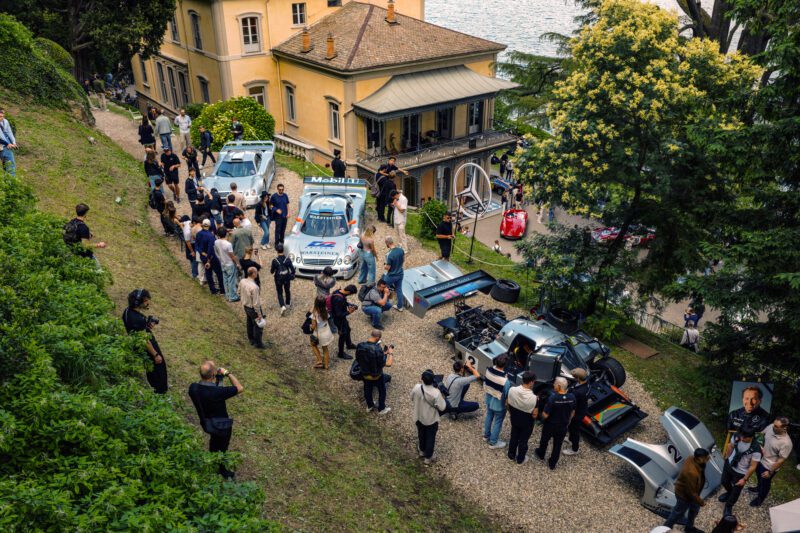 A group of people gathers around several vintage race cars, including a Mercedes-Benz CLK GTR, parked on a gravel path outside a large yellow villa near a lake at FuoriConcorso 2025, with trees and greenery surrounding the area.