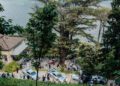 Aerial view of a lakeside villa surrounded by greenery, with several people and parked cars—including a sleek Mercedes-Benz—gathered near the entrance, set against hills across the water during FuoriConcorso 2025.
