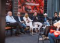 A panel of six people sits on a stage at FuoriConcorso 2025, discussing with microphones and drinks on the tables in front of them; an audience is visible in the foreground.
