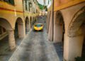 A yellow sports car cruises up a narrow, stone-paved street between old arches in a European town during the Ferrari Cavalcade in Siviglia, Andalusia.