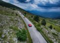 A group of Ferraris drives along a narrow mountain road in Andalusia, surrounded by rocky terrain and greenery under a cloudy sky, part of the thrilling Cavalcade Siviglia.