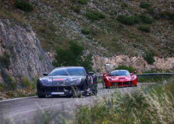 Two sports cars, a sleek black and a striking red Ferrari, race along a winding mountain road in Andalusia, bordered by rocky cliffs and lush greenery—the perfect scene for the exhilarating Cavalcade Siviglia.