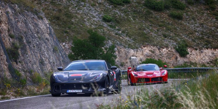 Two sports cars, a sleek black and a striking red Ferrari, race along a winding mountain road in Andalusia, bordered by rocky cliffs and lush greenery—the perfect scene for the exhilarating Cavalcade Siviglia.
