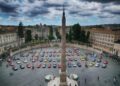 A tall stone obelisk stands at the center of a large plaza in Andalusia, lined with historic buildings, while rows of colorful Ferrari cars from the Cavalcade Siviglia event encircle the monument.