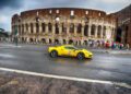 A yellow Ferrari speeds past the Colosseum in Rome on a wet road, with tourists gathered in the background, reminiscent of a Cavalcade Siviglia through historic landmarks.