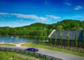 A purple Ferrari sports car drives on a paved road past a modern barn-style building, with a lake and wooded hills in the background under the blue skies of Andalusia.